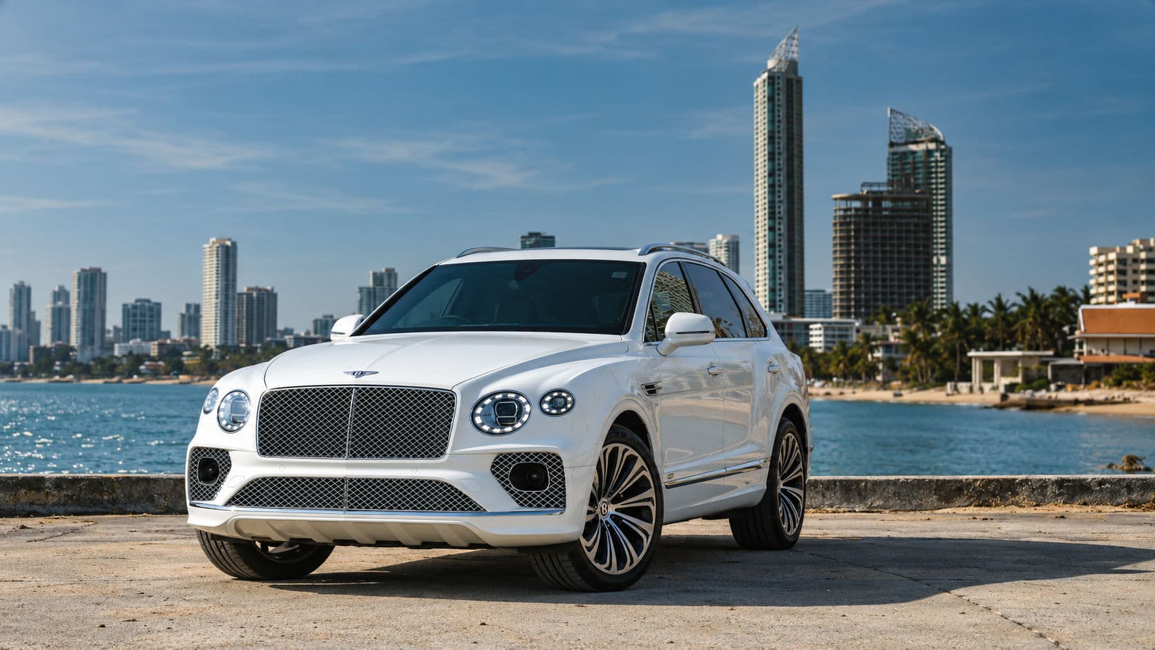 Bentayga V8 First Edition: White Bentley Bentayga V8 First Edition 2023, front three-quarter view by the sea with high-rise buildings in the background.