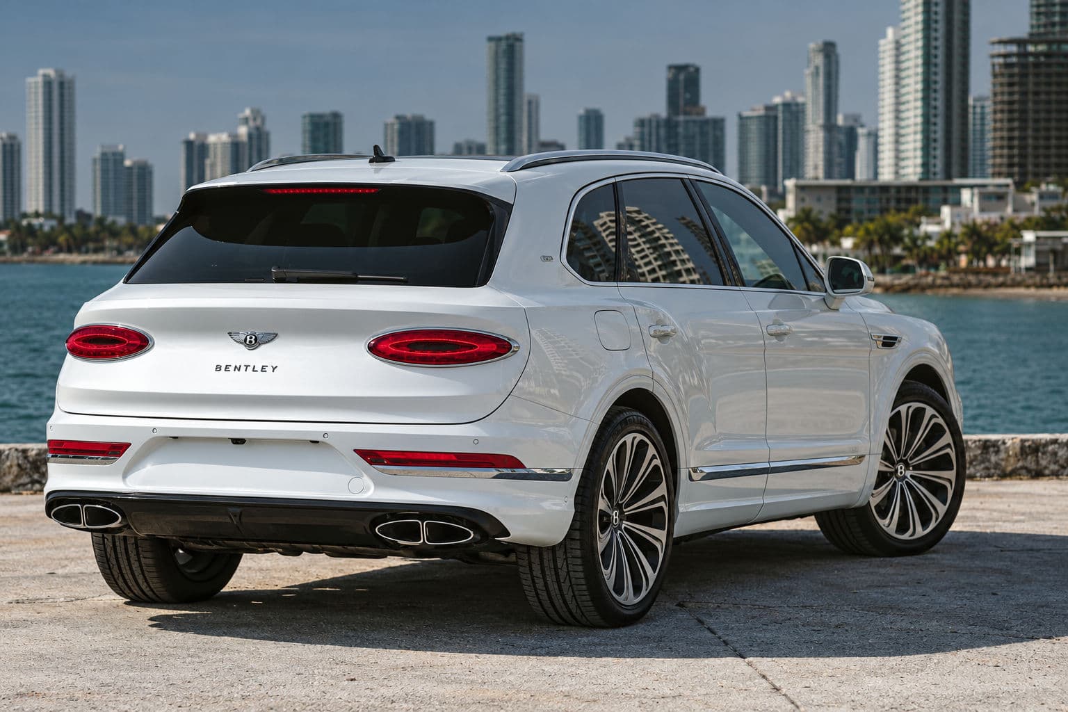 Bentayga V8 First Edition: White Bentley Bentayga V8 First Edition 2023, rear three-quarter view by the sea with high-rise buildings in the background.