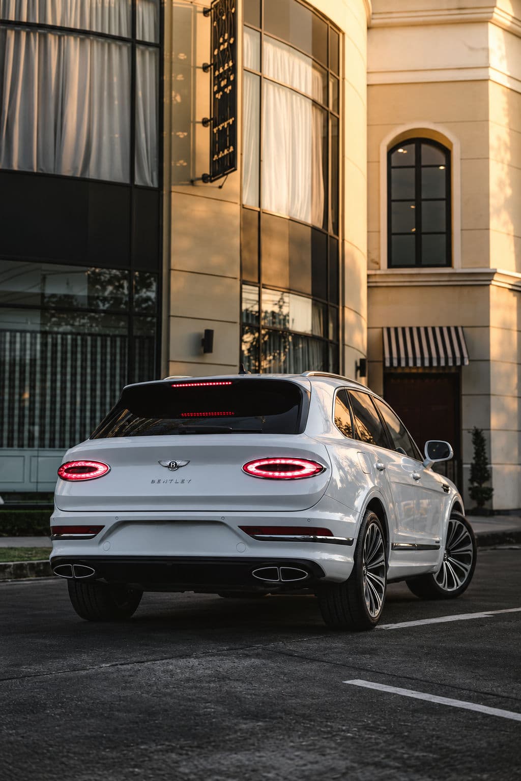 Bentayga V8 First Edition: White Bentley Bentayga V8 First Edition 2023, rear three-quarter view with illuminated taillights outside a building in warm evening light.