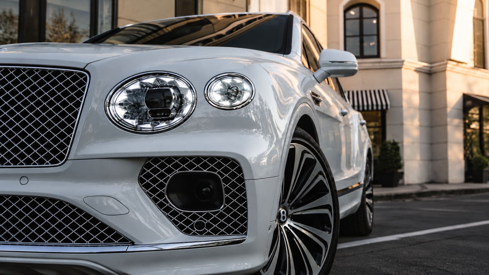 Bentayga V8 First Edition: White Bentley Bentayga V8 First Edition 2023, close-up of the front end showing the headlights, chrome mesh grille and front alloy wheel.
