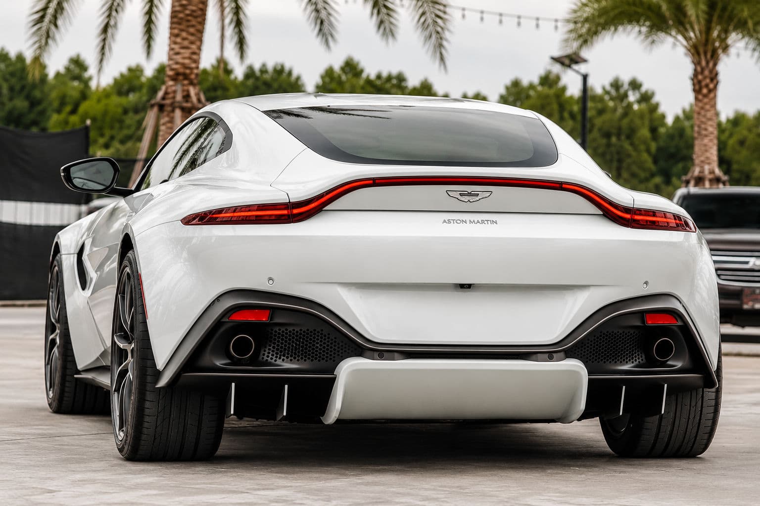 Vantage: White Aston Martin Vantage 2024 seen from a low rear angle, with illuminated red rear lights, a sculpted diffuser and twin round exhaust outlets against palm trees
