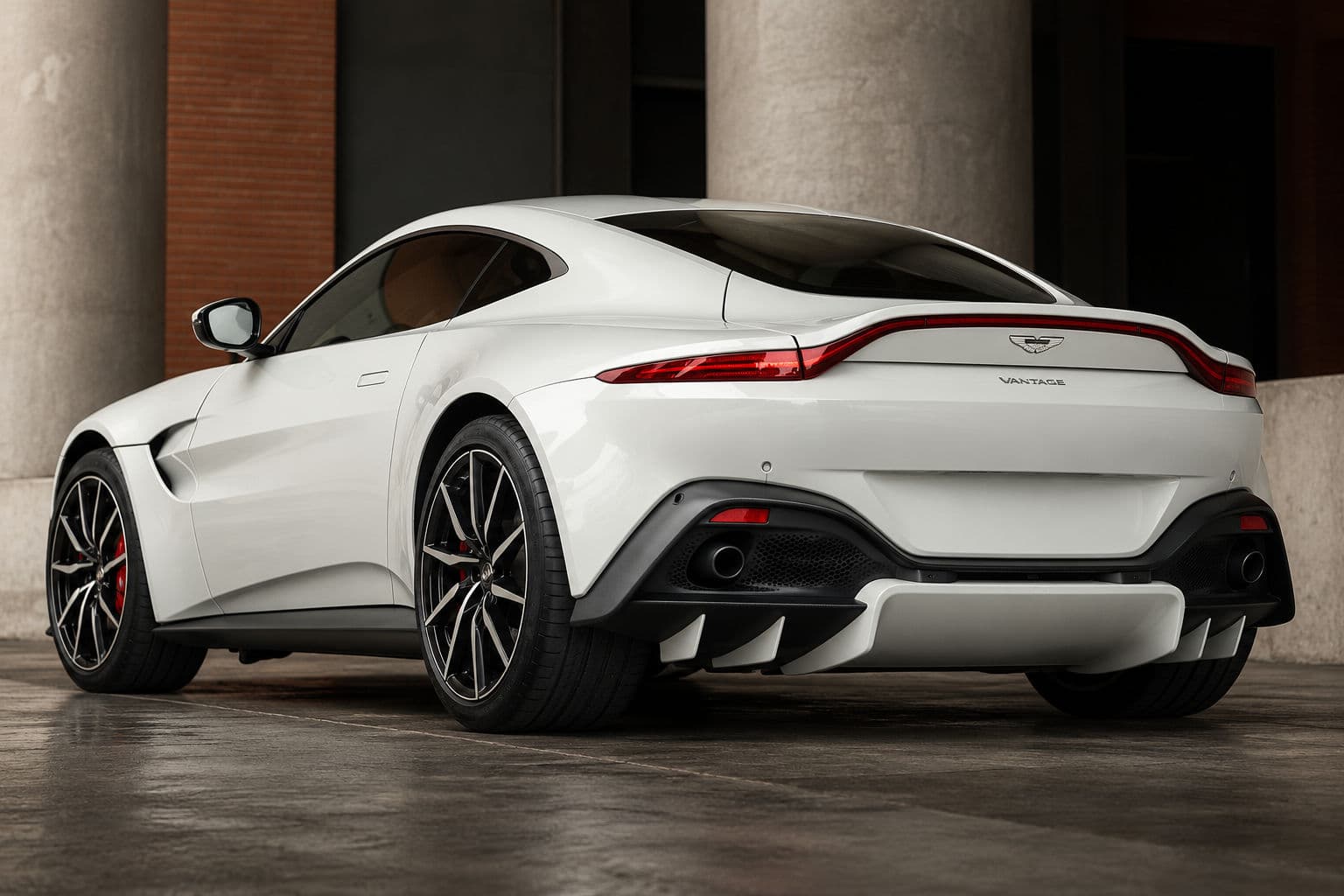 Vantage: White Aston Martin Vantage 2024 viewed from the rear left, showing the curved rear lights, diffuser and twin exhaust outlets against concrete columns and brickwork