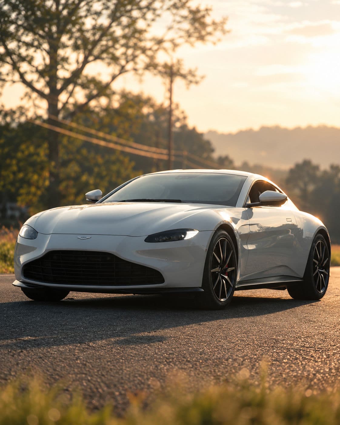 Vantage: White Aston Martin Vantage 2024 in a front three-quarter view on an asphalt road at sunset, with illuminated running lights and warm reflections across the body, backed by trees and distant hills