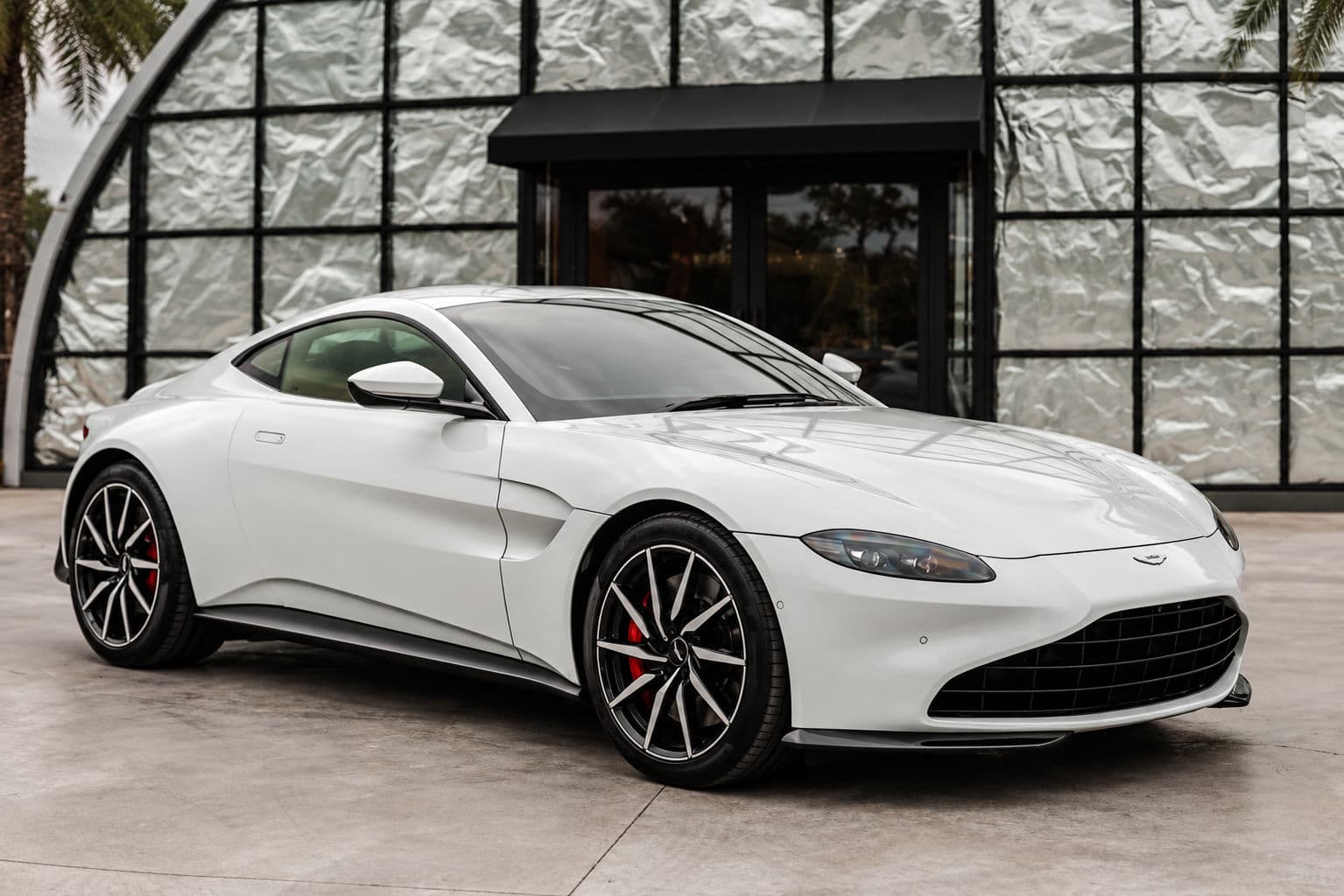 Vantage: White Aston Martin Vantage 2024 in a front three-quarter view, with a wide black grille, two-tone alloy wheels and red brake calipers outside a building with textured facade panels