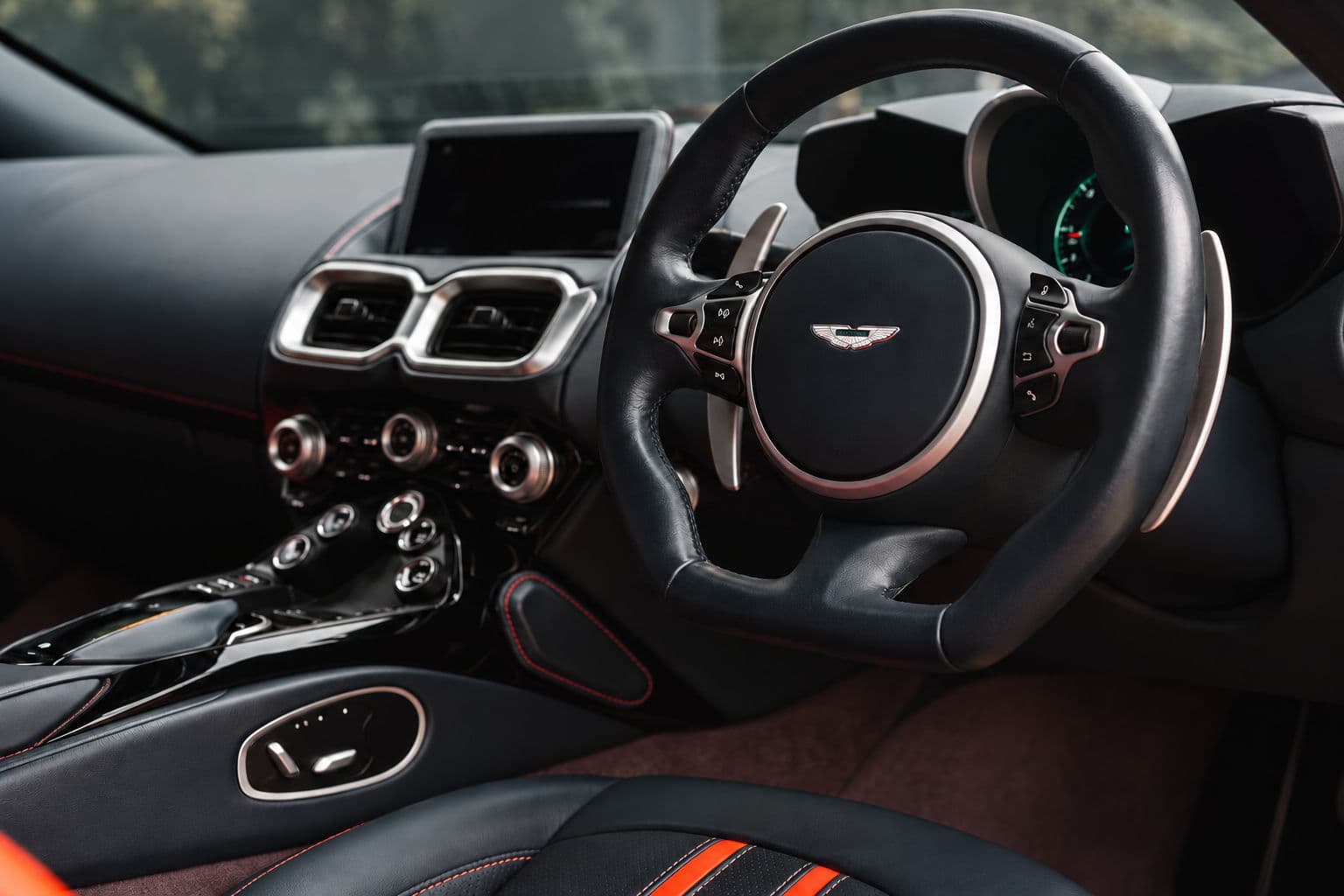 Vantage: Right-hand-drive interior of the Aston Martin Vantage 2024, showing a black leather steering wheel with paddle shifters, silver-trimmed console controls and a black seat with orange accents