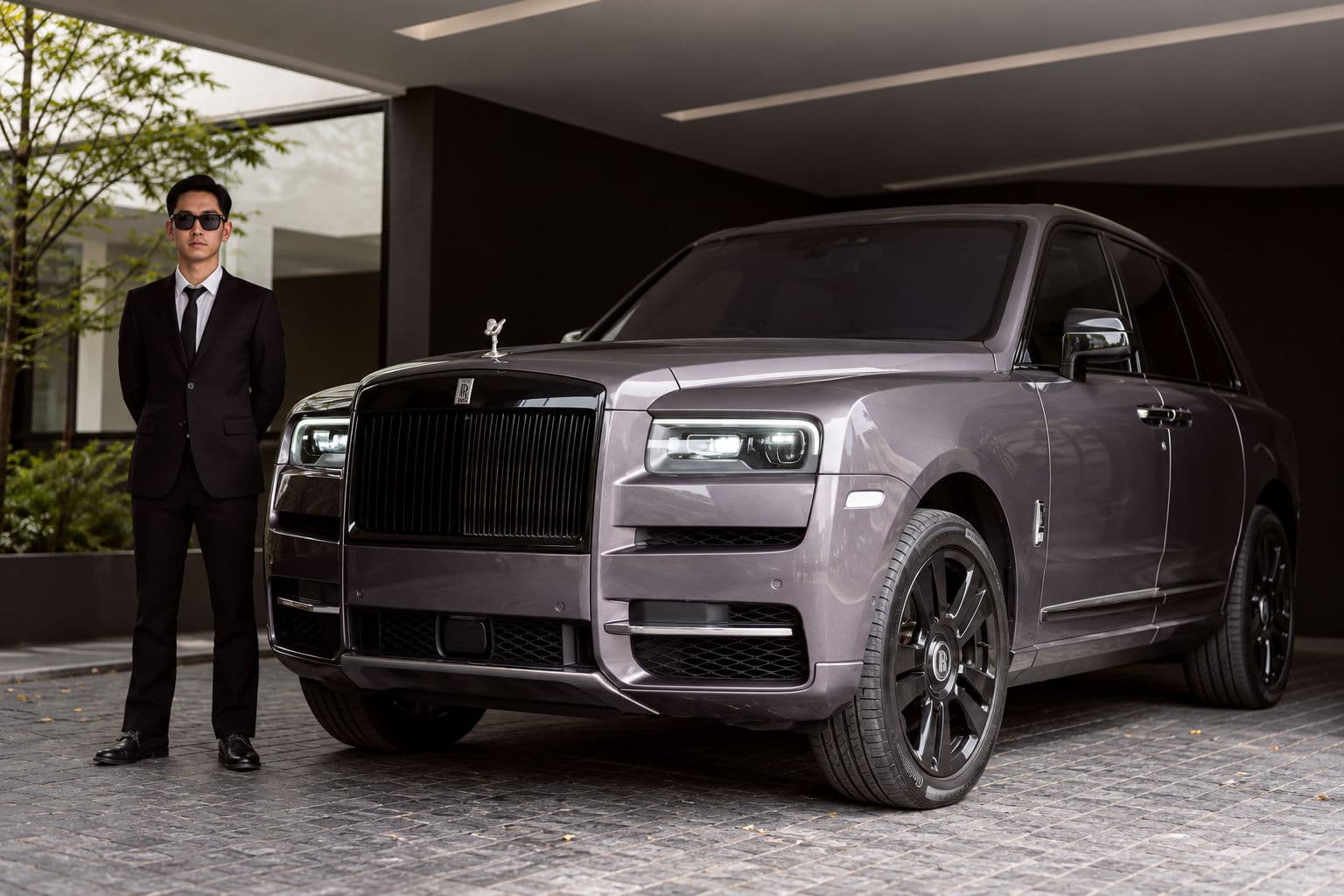 Cullinan: Rolls-Royce Cullinan 2024 in grey, front three-quarter view with chauffeur standing beside the car — RC24