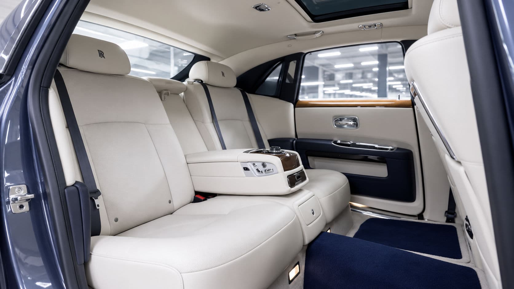 Ghost: Rolls-Royce Ghost 2019 rear cabin, two cream leather rear seats with RR headrest embroidery, dark blue carpet and centre armrest with wood trim, view through the open rear door
