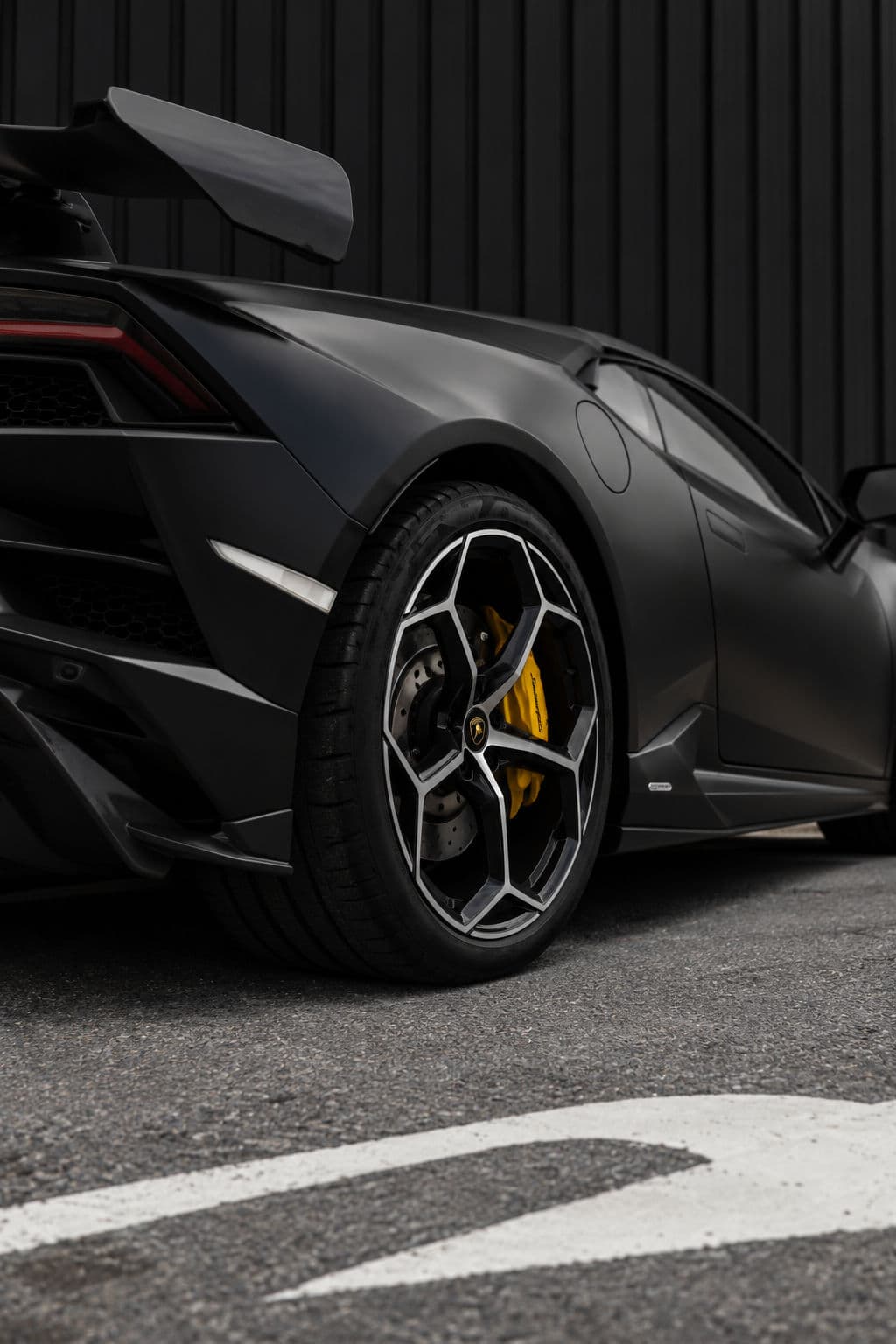 Huracan EVO RWD Spyder: Lamborghini Huracan EVO RWD Spyder 2023 in matte black, close-up of the rear wheel with a yellow brake caliper and the rear wing against a dark wall