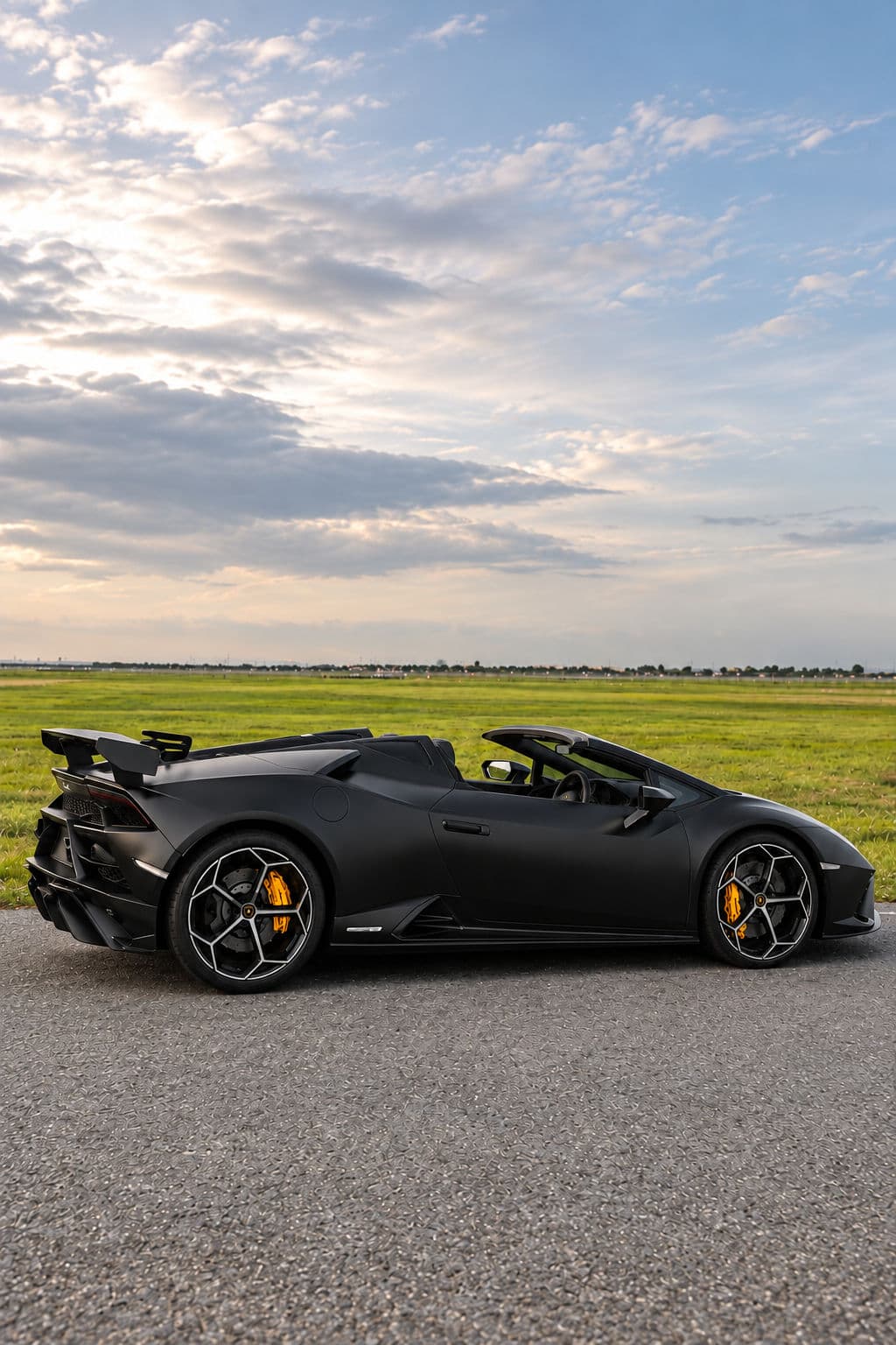 Huracan EVO RWD Spyder: Lamborghini Huracan EVO RWD Spyder 2023 in matte black with the roof down, side view against an open field and a cloudy sky